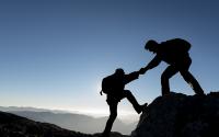 One climber offering a hand to another on top of a mountain with more mountains in the background